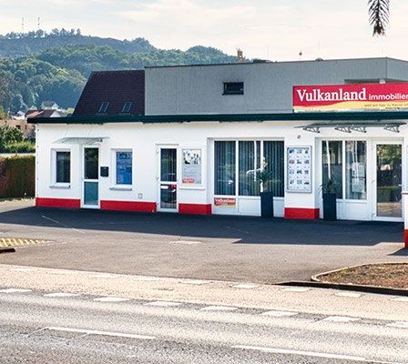 Ein Auto fährt an einem Geschäftsgebäude mit dem Namen 'Vulkanland Immobilien' vorbei. Das Gebäude hat Glasfenster, Türen und ein Schild. Ein silbernes Auto ist in der Nähe geparkt.