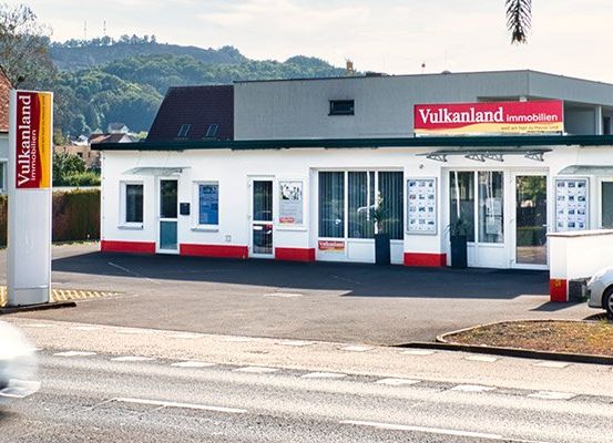 Ein Auto fährt an einem Geschäftsgebäude mit dem Namen 'Vulkanland Immobilien' vorbei. Das Gebäude hat Glasfenster, Türen und ein Schild. Ein silbernes Auto ist in der Nähe geparkt.