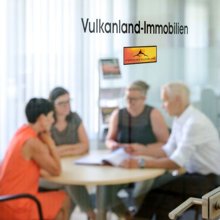 Vier Personen sitzen um einen Tisch in einem Büro. Die Glastür trägt den Schriftzug 'Vulkanland-Immobilien'.