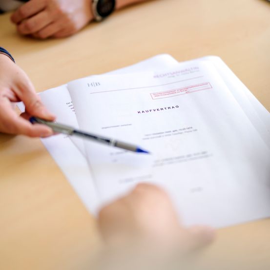 Eine Person unterzeichnet einen Kaufvertrag mit einem Stift auf einem Holztisch. Das Dokument ist in deutscher Sprache.
