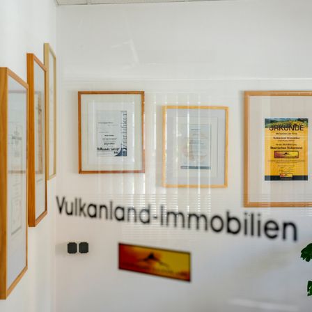 Ein Raum mit weißen Wänden, der gerahmte Zertifikate und Schilder zeigt. Das größte Schild trägt die Aufschrift 'Vulkanland-Immobilien'.
