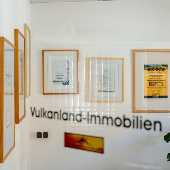 Ein Raum mit weißen Wänden, der gerahmte Zertifikate und Schilder zeigt. Das größte Schild trägt die Aufschrift 'Vulkanland-Immobilien'.