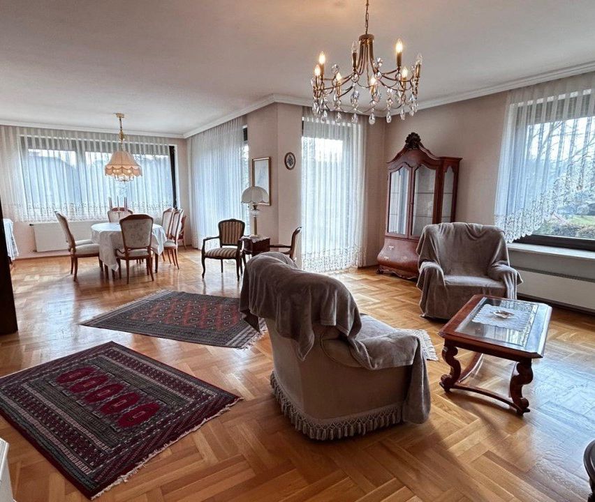 Ein Esszimmer mit Kronleuchter, Tisch und Stühlen. Es gibt zwei Teppiche auf dem Boden, einen hölzernen Schrank und einen Glastisch mit Lampe.
