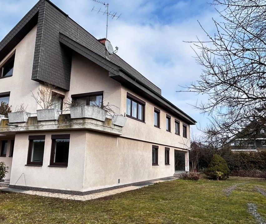Ein zweistöckiges Haus mit grauem Dach, mehreren Fenstern und einem Balkon mit Pflanzen auf der linken Seite.