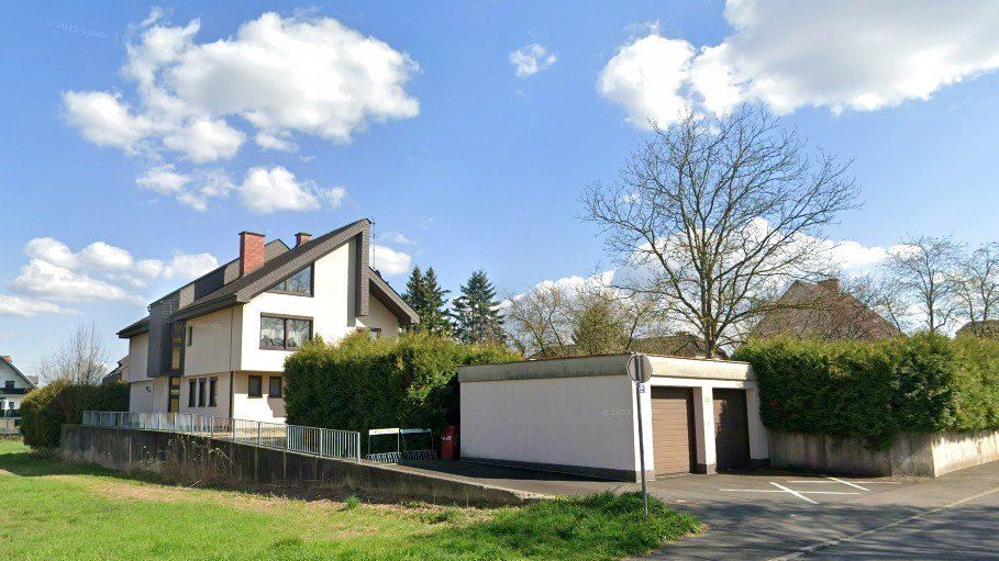 Ein zweistöckiges Haus mit Garage und Einfahrt, umgeben von Grün und einer Hecke, unter einem blauen Himmel mit Wolken.