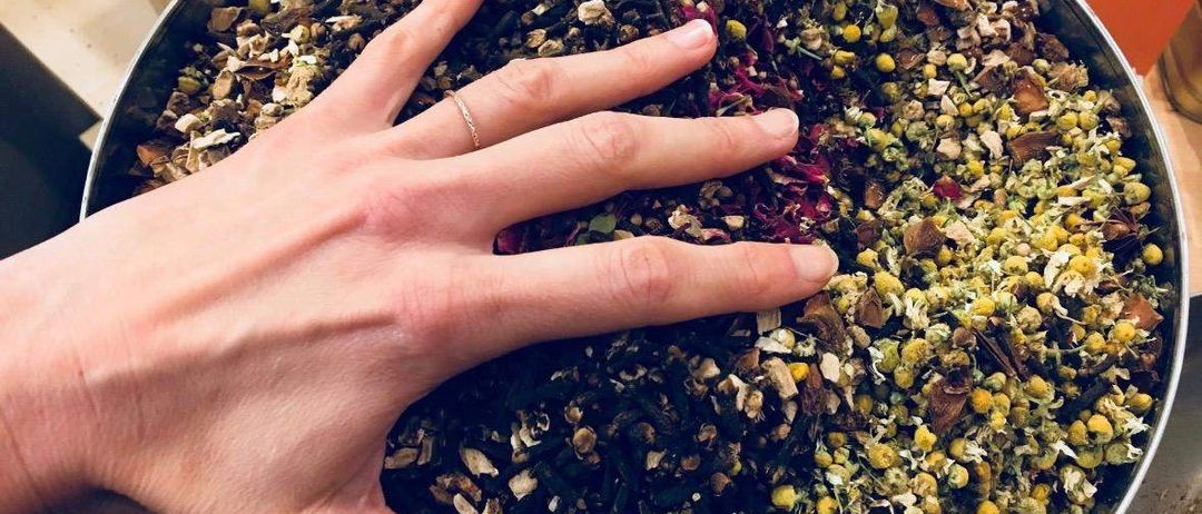 Eine Hand hält eine Schüssel mit getrockneten Kräutern, Gewürzen und Blumen. Die Kräuter sind eine Mischung aus verschiedenen Farben, und die Blumen sind rot und gelb. Die Schüssel ist aus Metall und steht auf einem Tisch.