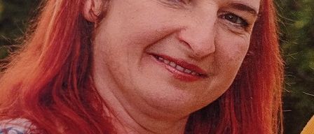 Eine Frau mit roten Haaren und einem Blumenhemd lächelt in die Kamera. Sie trägt eine Halskette mit bunten Perlen. Der Hintergrund ist verschwommen mit viel Grün.