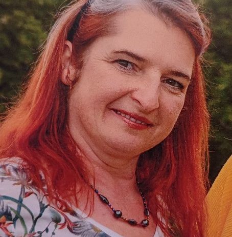 Eine Frau mit roten Haaren und einem Blumenhemd lächelt in die Kamera. Sie trägt eine Halskette mit bunten Perlen. Der Hintergrund ist verschwommen mit viel Grün.