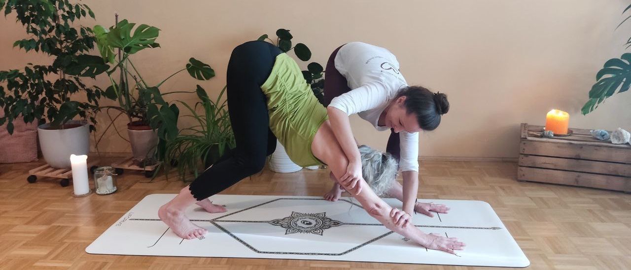 Eine Yoga-Instruktorin führt eine ältere Frau in eine Dehnungsposition auf einer Yogamatte. Im Hintergrund befinden sich Topfpflanzen und Kerzen.