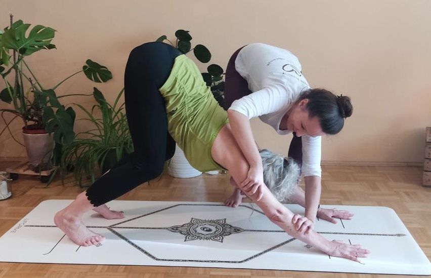 Eine Yoga-Instruktorin führt eine ältere Frau in eine Dehnungsposition auf einer Yogamatte. Im Hintergrund befinden sich Topfpflanzen und Kerzen.