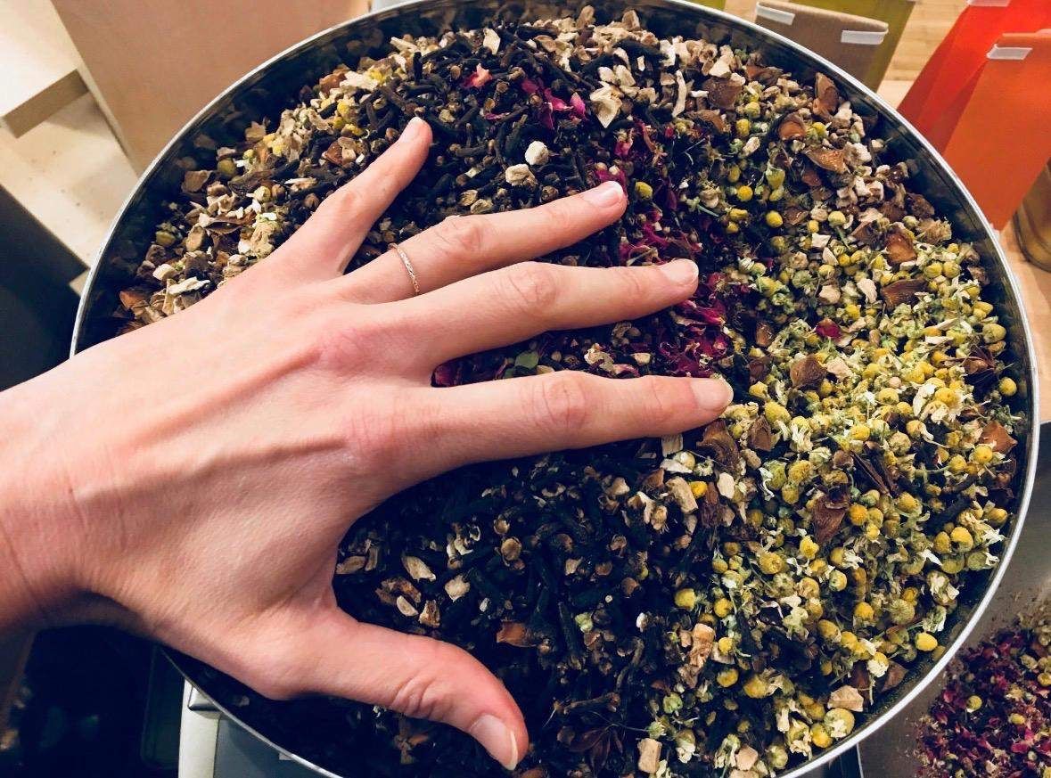 A hand gently touches a bowl full of various dried herbs and spices, including black tea, red petals, and yellow flowers.