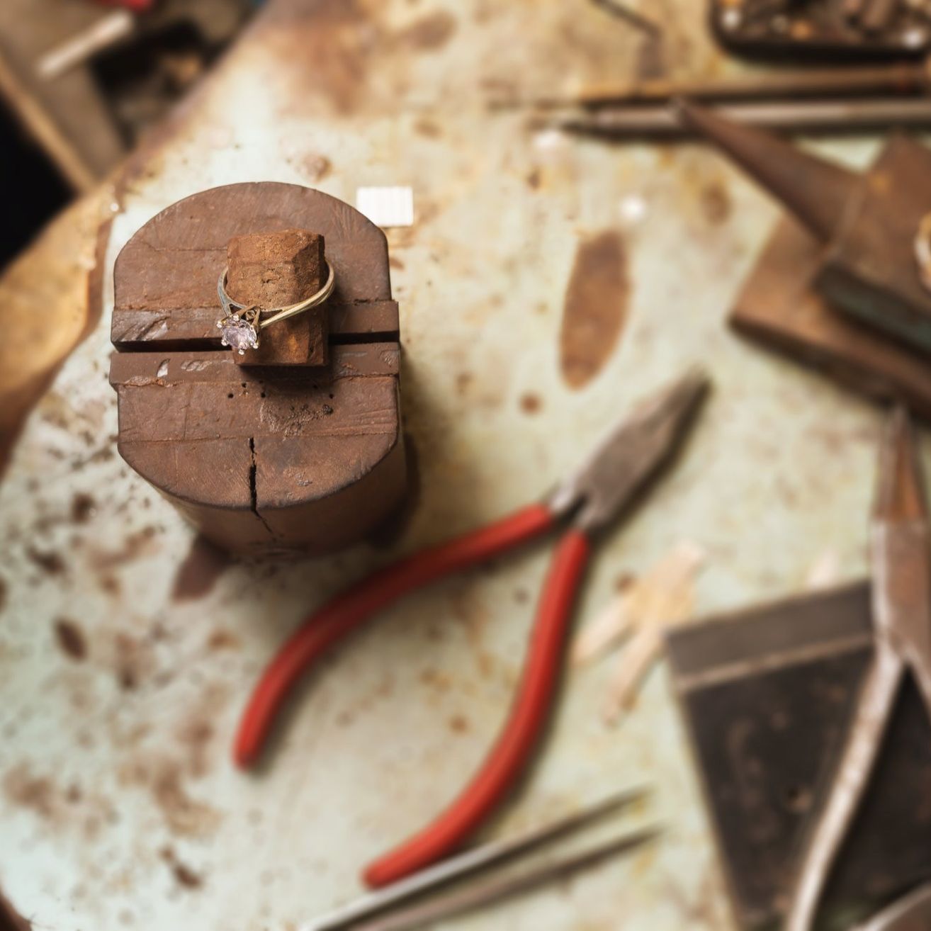 Ein Ring mit einem Diamanten ruht auf einem Holzblock auf einem überfüllten Arbeitstisch mit verschiedenen Werkzeugen, darunter Zangen und Meißeln.