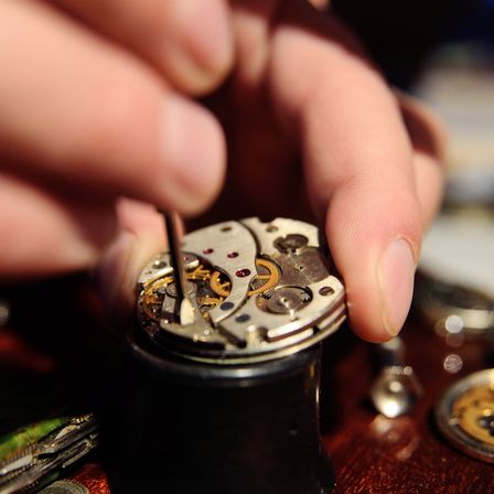 Eine Hand justiert das Innere eines Uhrwerks und konzentriert sich auf die komplexen Zahnräder. Der Hintergrund ist unscharf und zeigt einen Teil eines Holztisches.