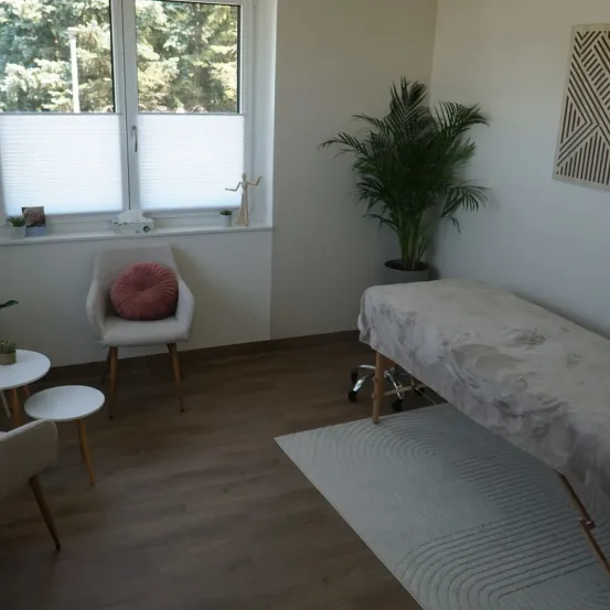 Ein Raum mit einem Fenster, einem Stuhl und einem Massagetisch. Eine Topfpflanze steht an der Wand und ein Teppich liegt auf dem Boden. Vor dem Fenster sind Bäume.