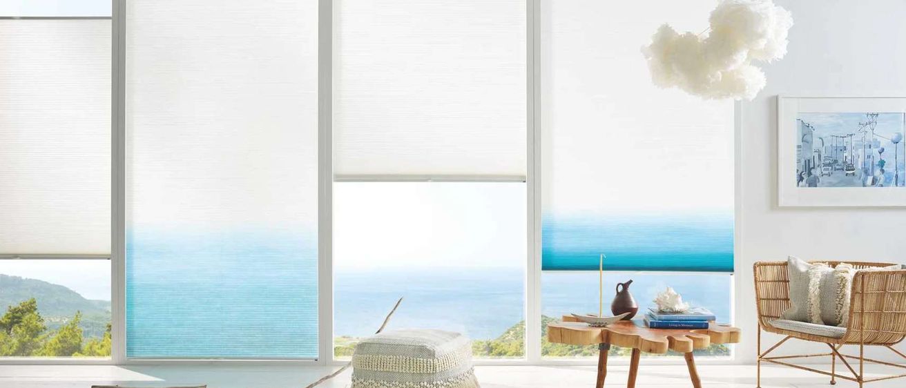 Ein Zimmer mit weißen Wänden und großen Fenstern, die einen Blick auf den Ozean zeigen. Ein weißer Hocker und ein Holztisch mit Büchern und einer Vase sind im Raum platziert.