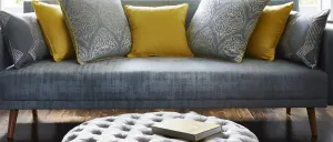 Ein graues Sofa mit einem vor dem Sofa stehenden, tuftigen Ottomanen, beide mit strukturierter Polsterung. Das Sofa hat dekorative Kissen in Blau und Gelb. Ein offenes Buch liegt auf dem Ottomanen.