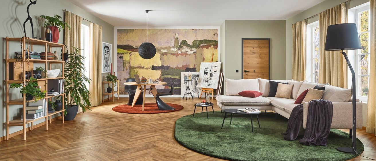 Ein modernes Wohnzimmer mit Holzboden, einem großen Gemälde, einem weißen Sofa mit verschiedenen Kissen, einem runden grünen Teppich, einer Holztür und einem Tisch mit Stühlen.