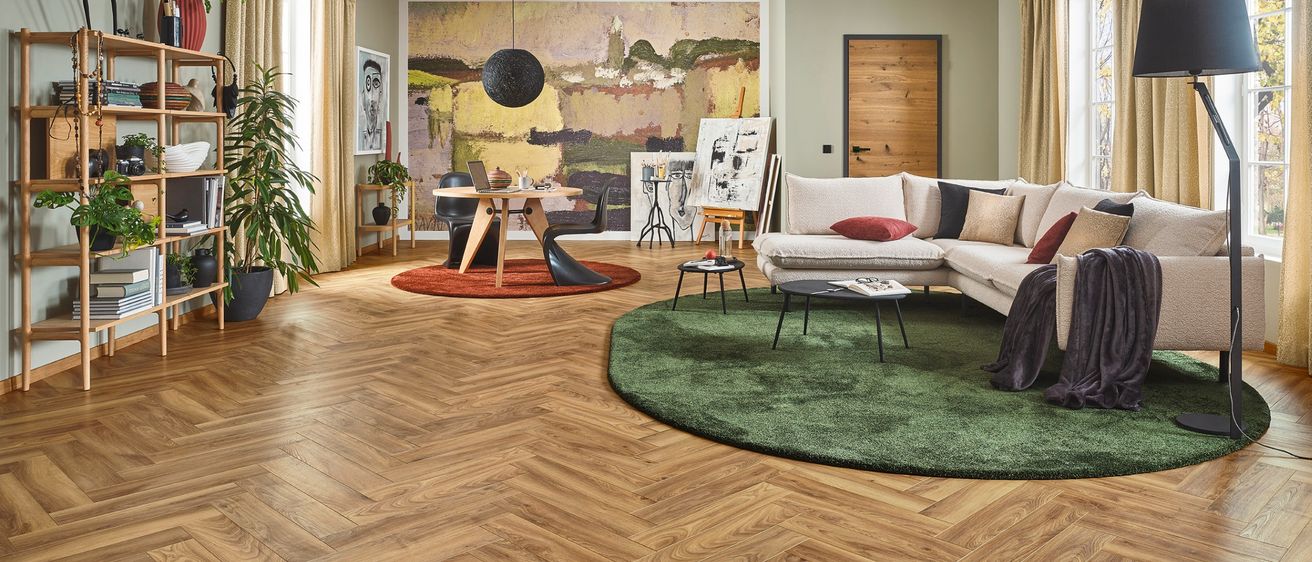 Ein modernes Wohnzimmer mit Holzboden, einem großen Gemälde, einem weißen Sofa mit verschiedenen Kissen, einem runden grünen Teppich, einer Holztür und einem Tisch mit Stühlen.