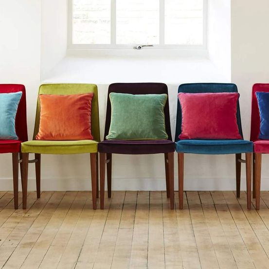 Fünf Stühle mit farbenfrohen Kissen sind in einer Reihe vor einer weißen Wand mit einem Fenster angeordnet. Der Boden ist aus Holz.