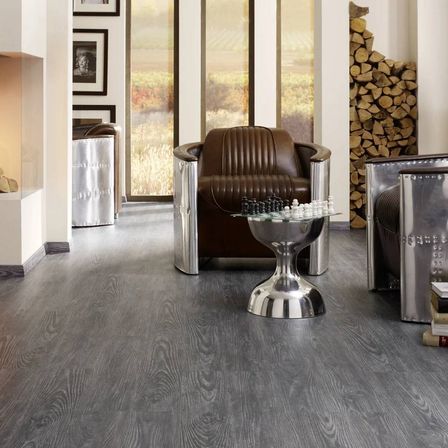 Ein Innenraum mit einem grauen Holzboden, einem braunen Ledersessel und einem Glastisch mit Schachfiguren. Es gibt gerahmte Bilder an der Wand und einen Kamin.