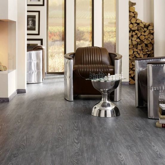 Ein Innenraum mit einem grauen Holzboden, einem braunen Ledersessel und einem Glastisch mit Schachfiguren. Es gibt gerahmte Bilder an der Wand und einen Kamin.
