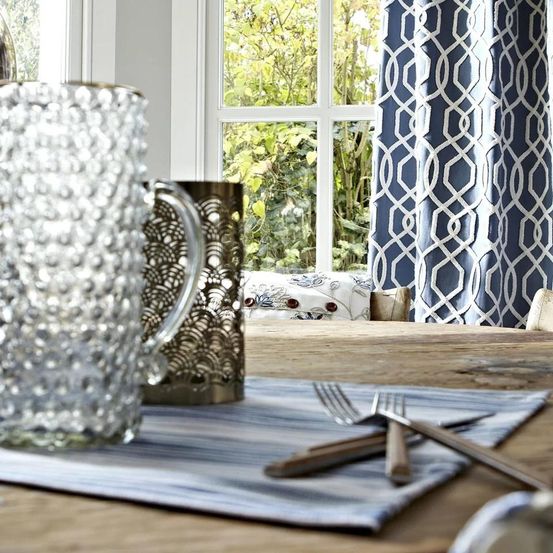 Ein Esstisch mit einer blau-weißen Tischdecke, einer Glaskanne, einem Metallbecher, Gabeln und Messern. Der Tisch hat ein Fenster mit weißen und blauen Vorhängen.