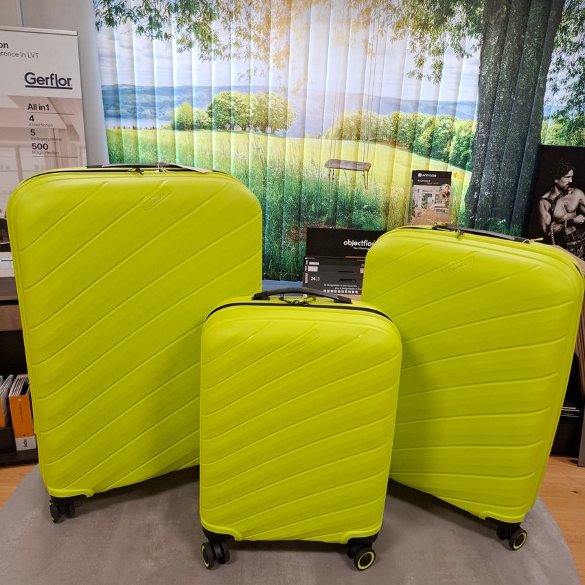 Drei hellgrüne Koffer mit Rädern auf einer grauen Oberfläche. Dahinter ist eine Kulisse mit einer grünen Landschaft mit Bäumen und einem Mann in einem schwarzen T-Shirt.