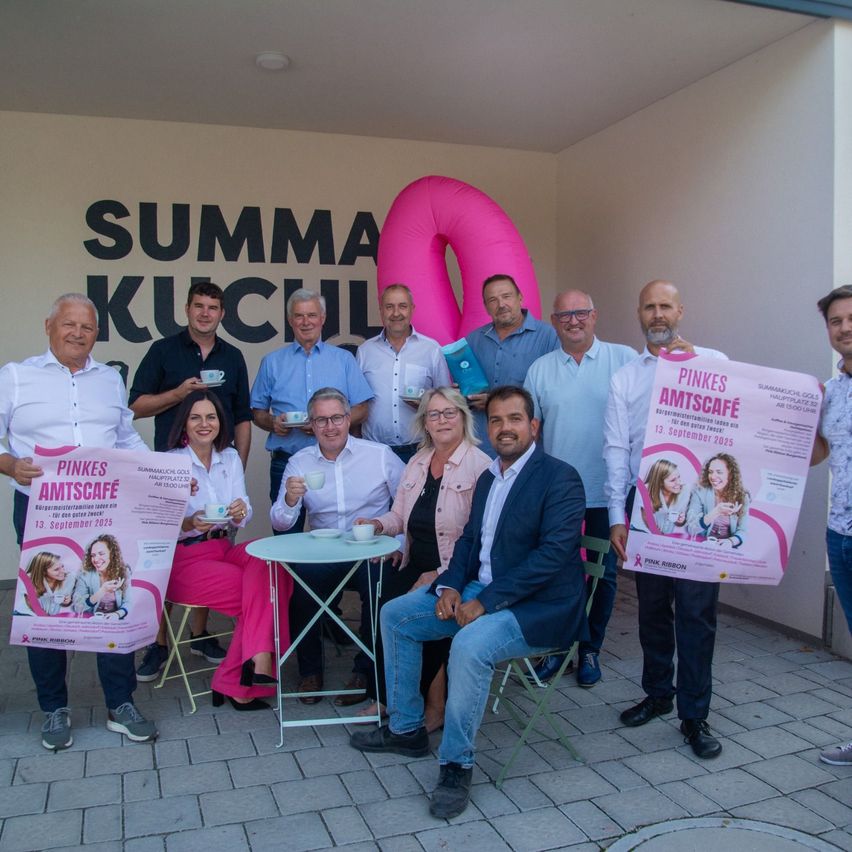 Eine Gruppe von Menschen steht vor einem rosa Luftballon und einem Banner. Sie halten rosa Plakate und trinken Kaffee. Die Banner tragen den Text 'Pinkes Amtescafe' und das Datum '13. September 2025'.