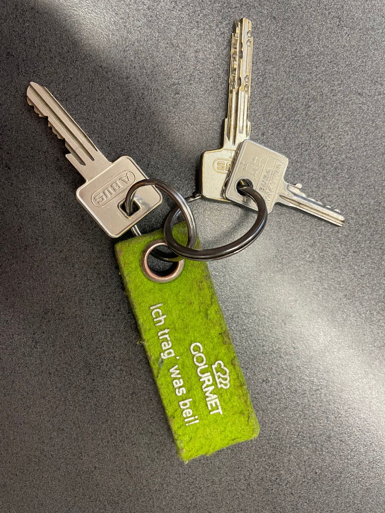 Ein Schlüsselanhänger mit zwei silbernen Schlüsseln und einem grünen Schlüsselanhänger, der 'Ich trage was bei!' und 'COURMET' liest, liegt auf einer grauen Oberfläche.