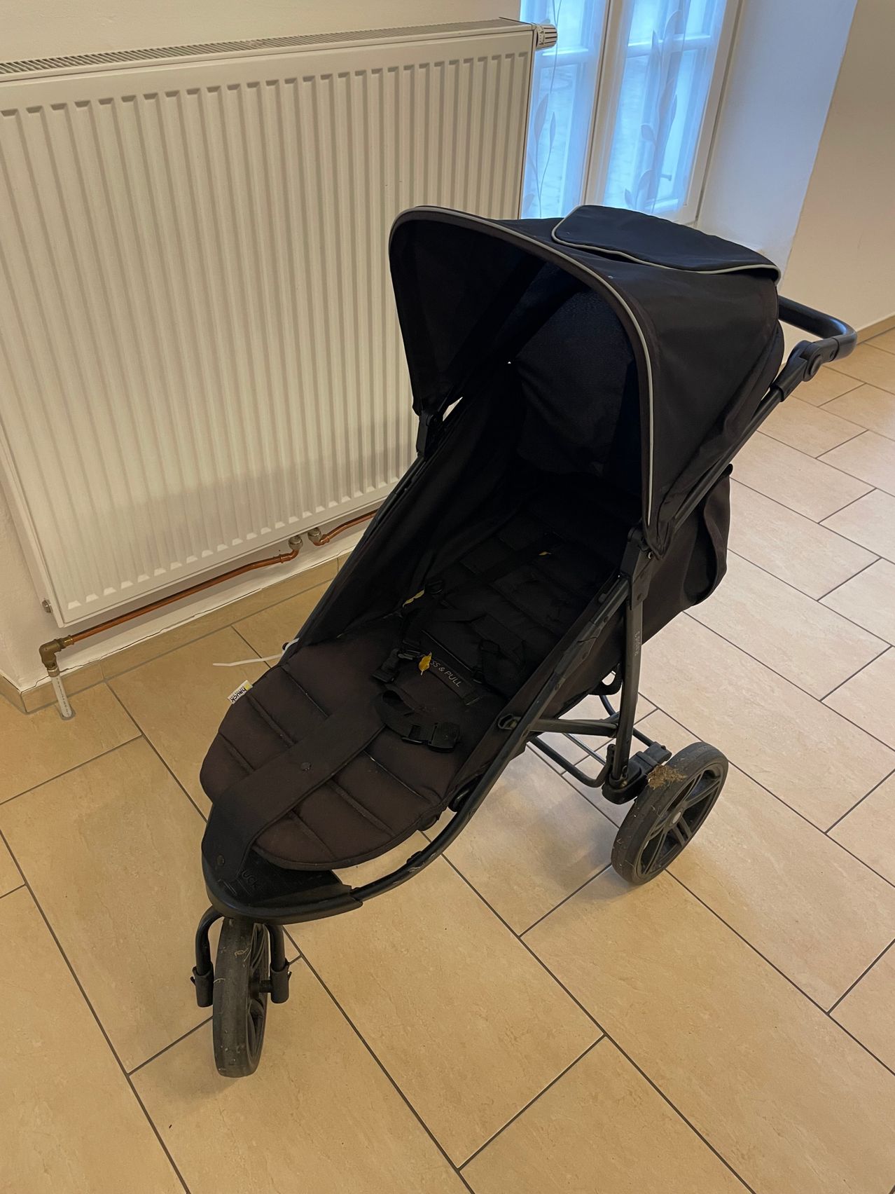 Ein schwarzer Kinderwagen mit schwarzem Sitz, Verdeck und Rädern steht auf einem gefliesten Boden mit einem weißen Heizkörper im Hintergrund.