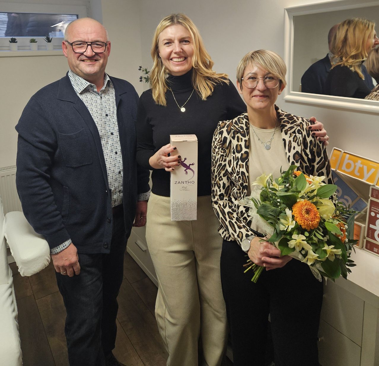 Drei Personen stehen eng beieinander in einem Raum. Ein Mann und eine Frau halten eine Schachtel mit einem Eidechsenaufdruck und Blumen, während eine Frau lächelt. Hinter ihnen reflektiert ein Spiegel eine Frau.