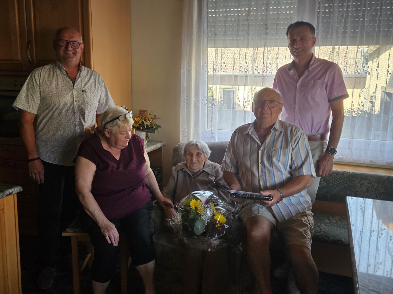 Fünf Menschen posieren für ein Foto im Wohnzimmer. Eine ältere Frau sitzt auf einem Sofa mit einem Blumenstrauß. Der Mann neben ihr hält ein Buch. Hinter ihnen stehen zwei Männer und eine Frau.