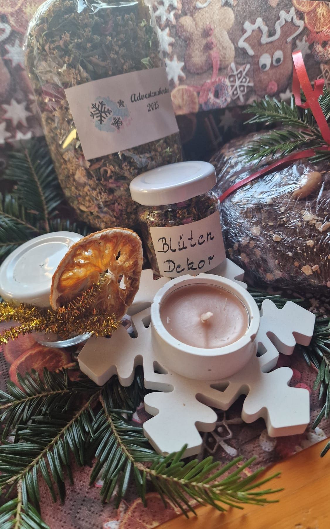 Ein festlicher Tisch zeigt eine Kerze, eine getrocknete Orangenscheibe und eine schneeketenförmige Platte, umgeben von Tannenzweigen, einem eingewickelten Geschenk und Gläsern mit Kräutern und Blumendekoration.