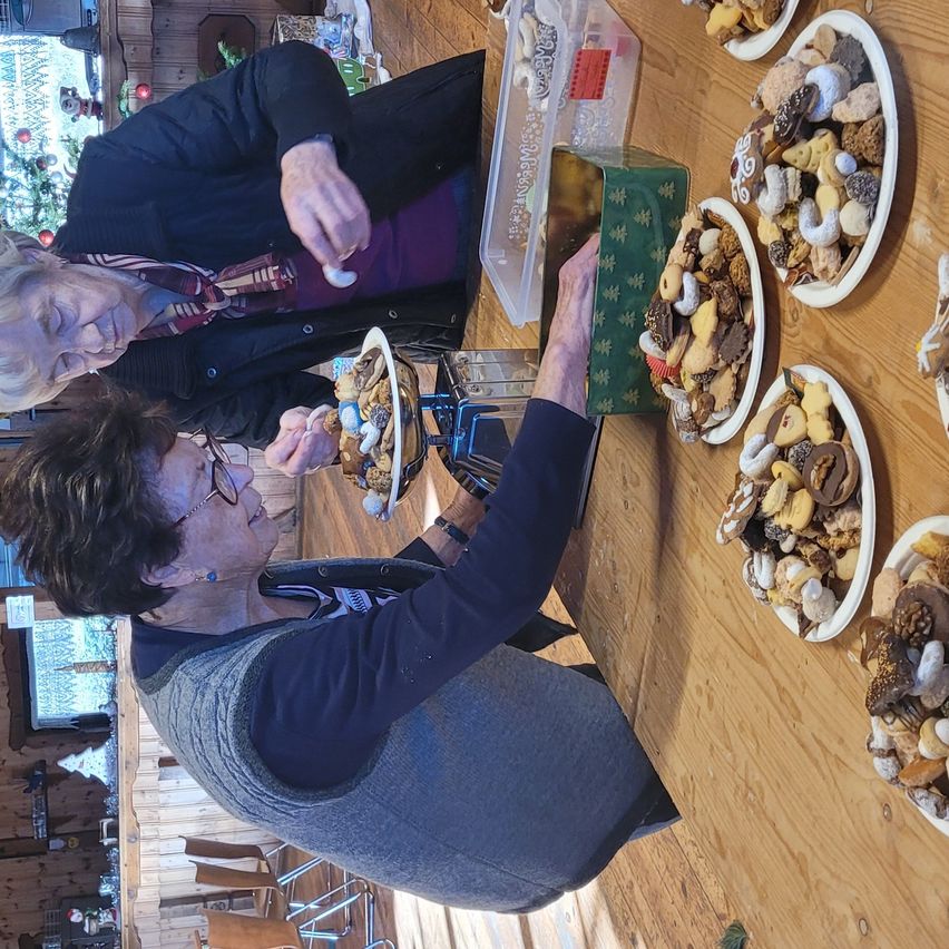 Zwei Frauen genießen einen Teller mit Keksen in einer festlichen Umgebung. Ein Tisch ist mit verschiedenen Platten gefüllt mit Keksen.