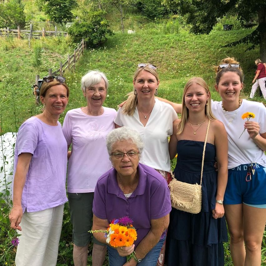 Eine Gruppe von Frauen, einschließlich einer älteren Frau mit Blumen, steht in einem Garten mit einem grasbewachsenen Hügel im Hintergrund.