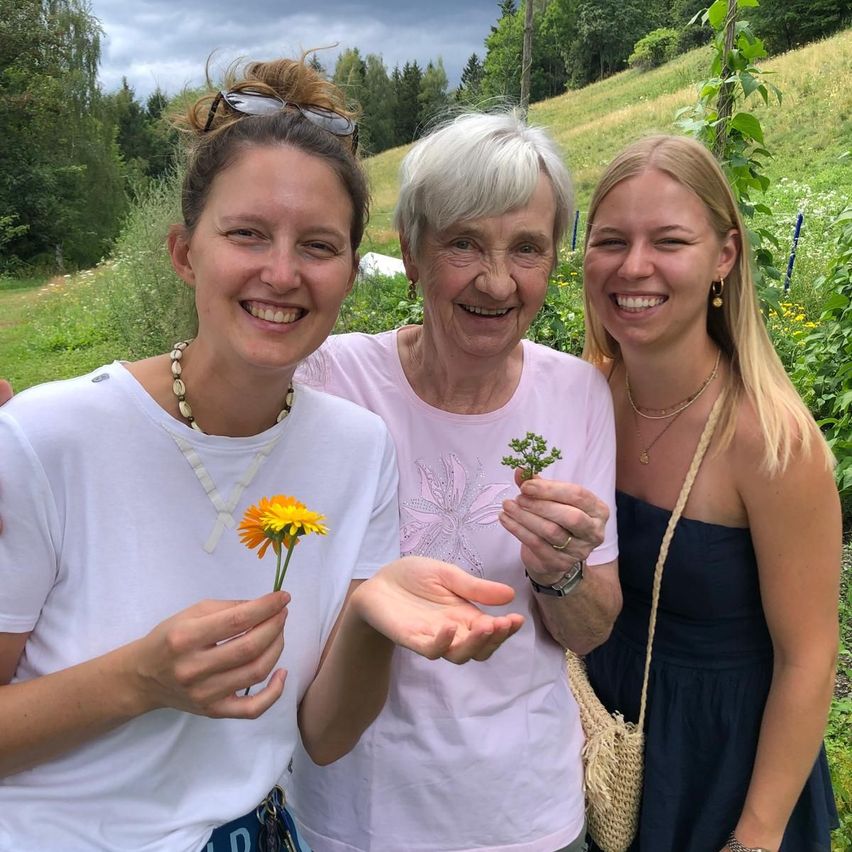 Drei Frauen in einem Garten, zwei halten Blumen, lächeln und posieren für ein Foto. Die Frau auf der linken Seite trägt ein weißes Shirt und eine Halskette, die mittlere Frau ein rosa Shirt und die rechte Frau ein blaues Kleid.