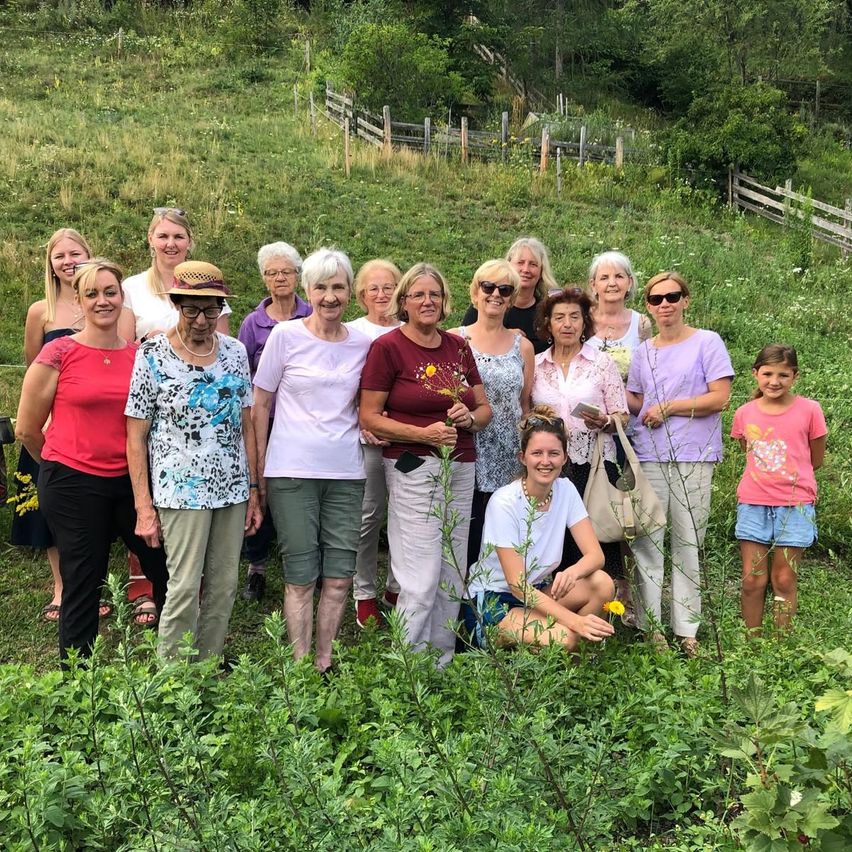 Eine Gruppe von Frauen und einem jungen Mädchen steht in einem Feld mit hohem Gras und Blumen. Sie lächeln und scheinen für ein Foto zu posieren.