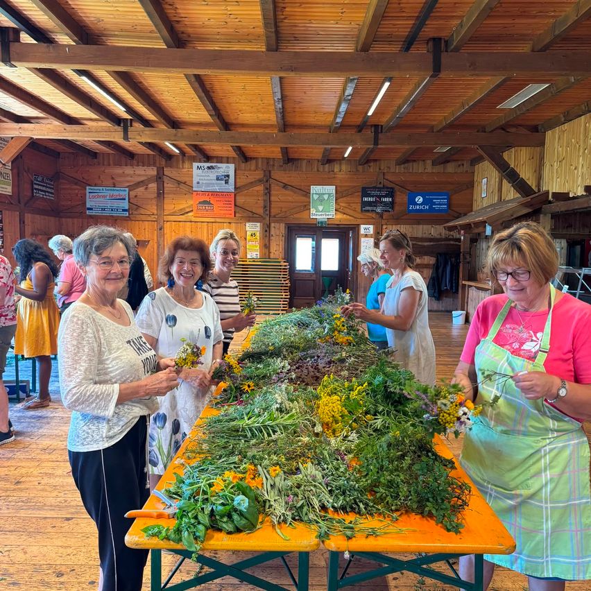 A group of women in aprons are standing around a table filled with herbs and flowers, possibly in a workshop.