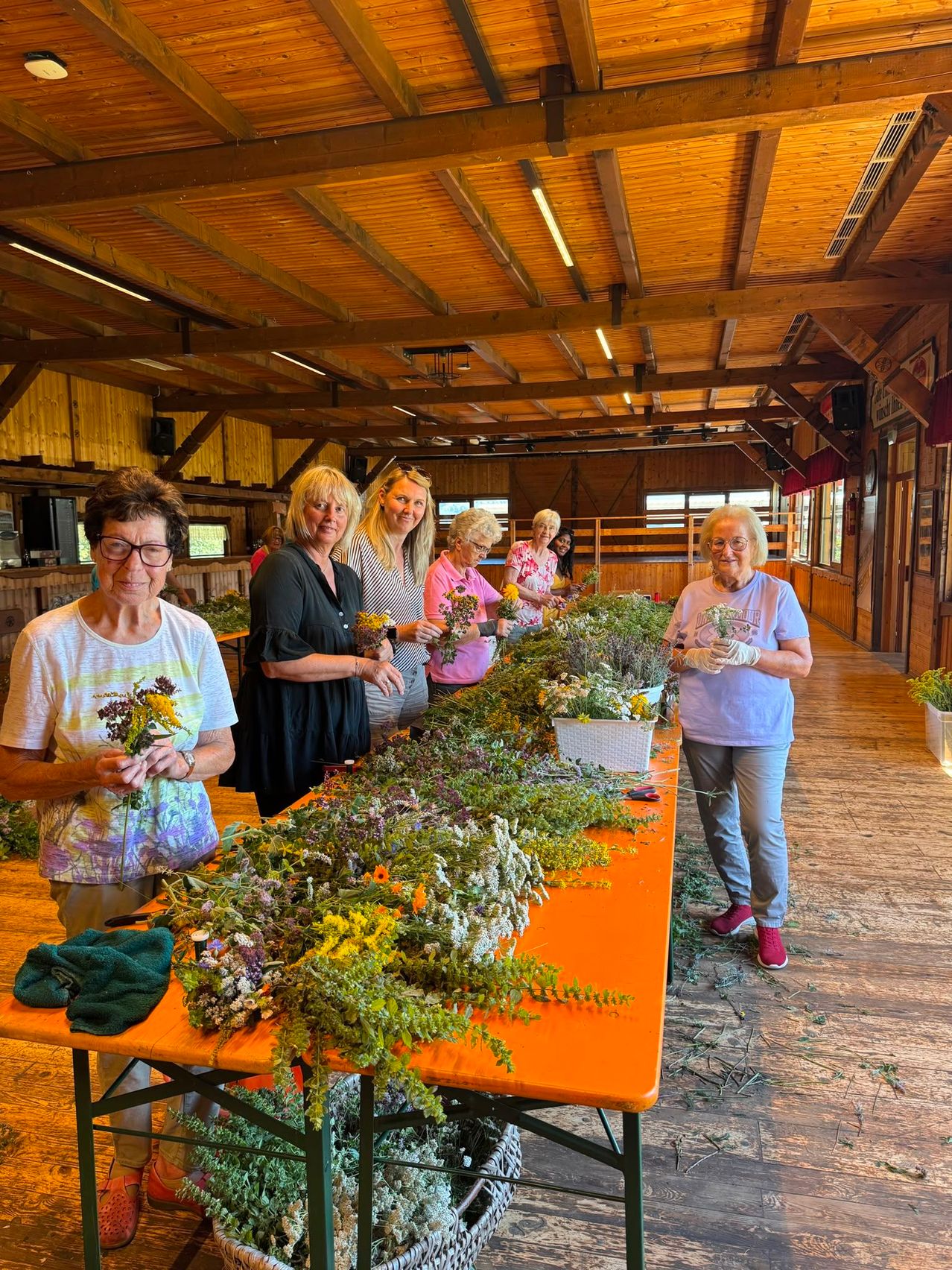 A group of women are gathered around a long table filled with various flowers and plants, possibly in a workshop setting. They are holding flowers and seem to be working on them. The room has wooden walls and a ceiling.