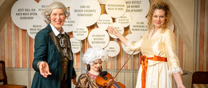 Drei Personen in Kostümen aus der Vergangenheit. Ein Mann, eine Frau und ein junges Mädchen mit einer Geige. Sie stehen vor einer Wand mit runden Aufklebern mit deutschen Phrasen.
