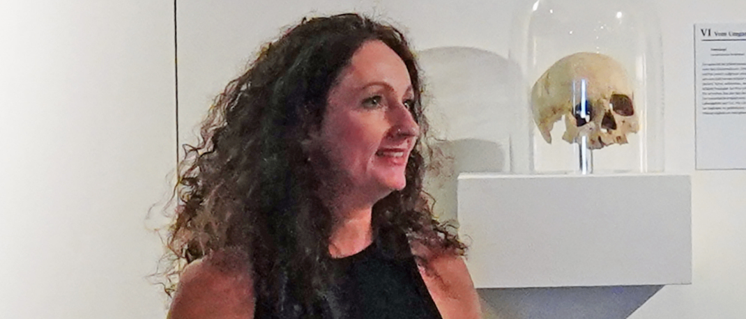 Eine Frau steht in einem Museum und schaut sich einen Schädel in einem Glaskasten an. Der Text lautet 'Museumsabend von Perchten, Hexen und Vampiren'. Vortrag von Mag. Elke Ferderbar, Landesmuseum Burgenland. Donnerstag, 16. Oktober 2025 um 18 Uhr.