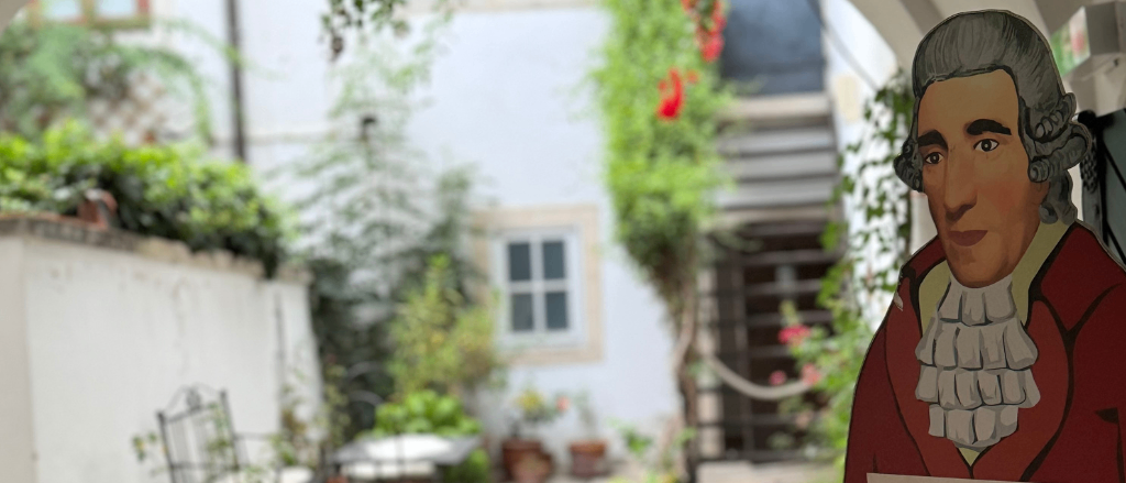 Ein Mann steht neben einem Schild, das den Eingang anzeigt, mit einem Innenhof voller Pflanzen und einem Tisch dahinter.