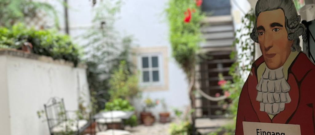 Ein Innenhof mit einer Statue eines Mannes, der ein Schild mit der Aufschrift 'Eingang' hält. Hinter ihm befindet sich eine Treppe und eine weiße Wand. Im Vordergrund stehen Topfpflanzen und ein Tisch mit Stühlen.