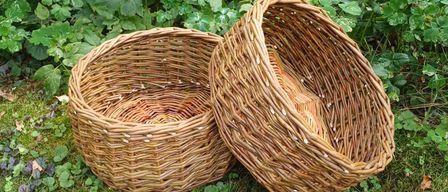 Zwei Weidenkörbe stehen auf Gras mit grünen Pflanzen im Hintergrund. Ein Korb ist leer, während der andere Gegenstände enthält.