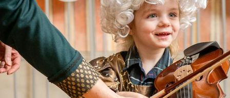Ein lächelndes Kind mit lockigem weißem Haar spielt Geige. Eine Hand hält den Hals der Geige, vermutlich beim Unterrichten. Das Kind trägt ein farbenfrohes Kostüm. Der Hintergrund ist verschwommen.