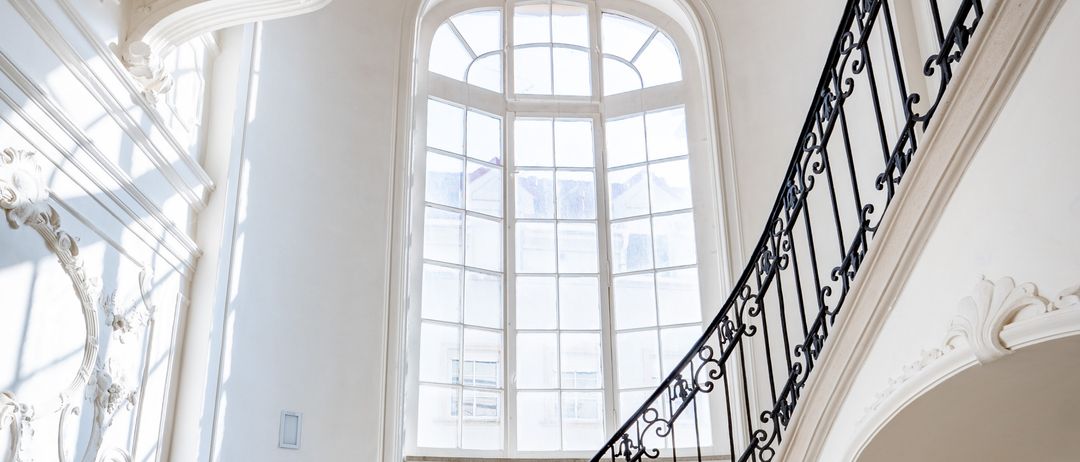 Eine verzierte Treppe mit schwarzem Schmiedeeisengeländer, Marmorstufen und weißen Wänden. Oben hängt eine Kronleuchter von der Decke. Ein großes bogenförmiges Fenster mit Glasscheiben ist sichtbar.