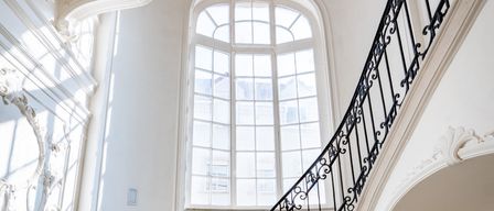 Eine verzierte Treppe mit schwarzem Schmiedeeisengeländer, Marmorstufen und weißen Wänden. Oben hängt eine Kronleuchter von der Decke. Ein großes bogenförmiges Fenster mit Glasscheiben ist sichtbar.