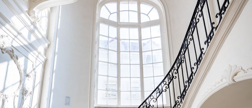 Eine verzierte Treppe mit schwarzem Schmiedeeisengeländer, Marmorstufen und weißen Wänden. Oben hängt eine Kronleuchter von der Decke. Ein großes bogenförmiges Fenster mit Glasscheiben ist sichtbar.