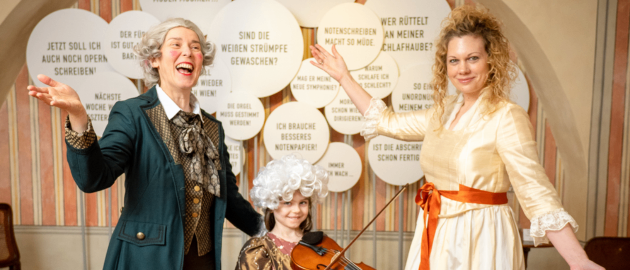 Drei Personen in historischer Kleidung stehen zusammen. Eine Frau in einem grünen Mantel zeigt mit ihren Händen. Ein junges Mädchen mit Perücke hält eine Geige. Eine Frau in einem gelben Kleid hält ein Band.