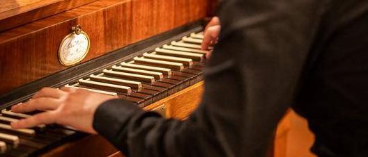 Ein Mann spielt ein Holzklavier in einem schwach beleuchteten Raum. Er sitzt auf einem Stuhl, mit zwei Personen hinter ihm. Das Klavier hat einen goldenen Knopf und Tasten.